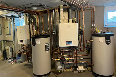 Two wall-hung boilers above two white indirect hot water tanks, joined by copper piping in a basement