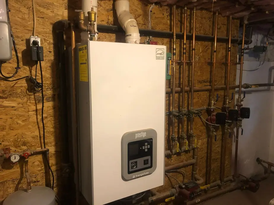 A wall-hung boiler in a basement with black-iron and copper piping and zone valves on a plywood panel