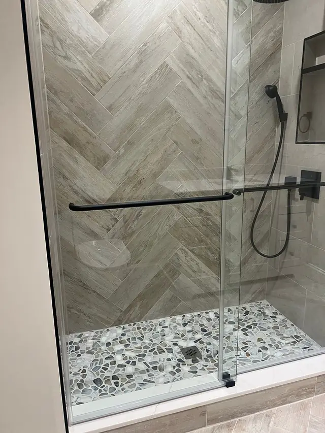 A finished walk-in shower with herringbone wall tile, a pebble floor, black rain head and fixtures behind a sliding glass door
